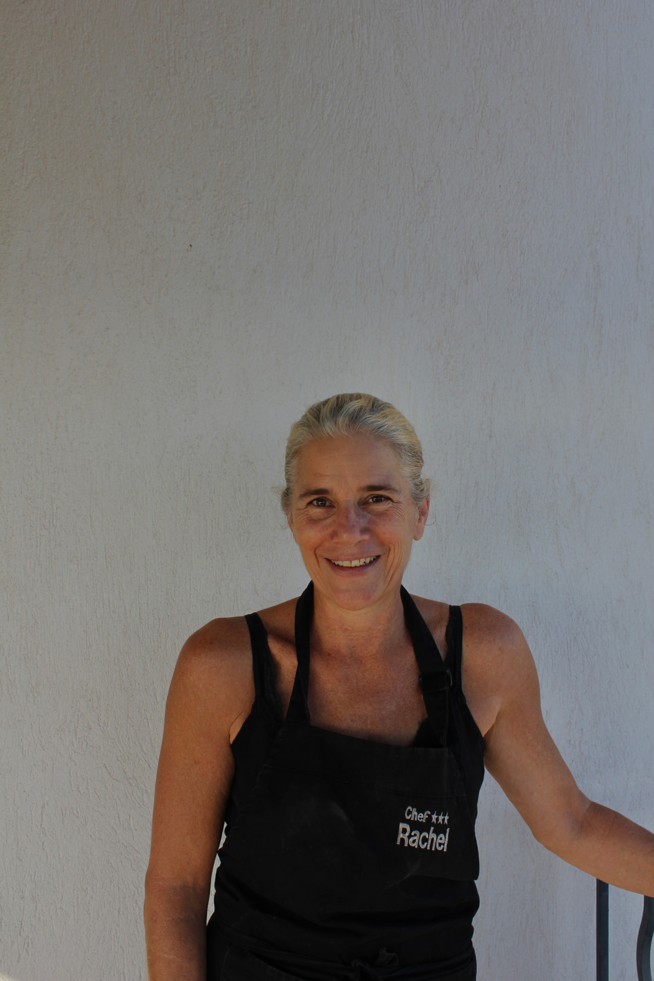 Chef Rachel souriante, en tablier noir brodé, devant un mur clair